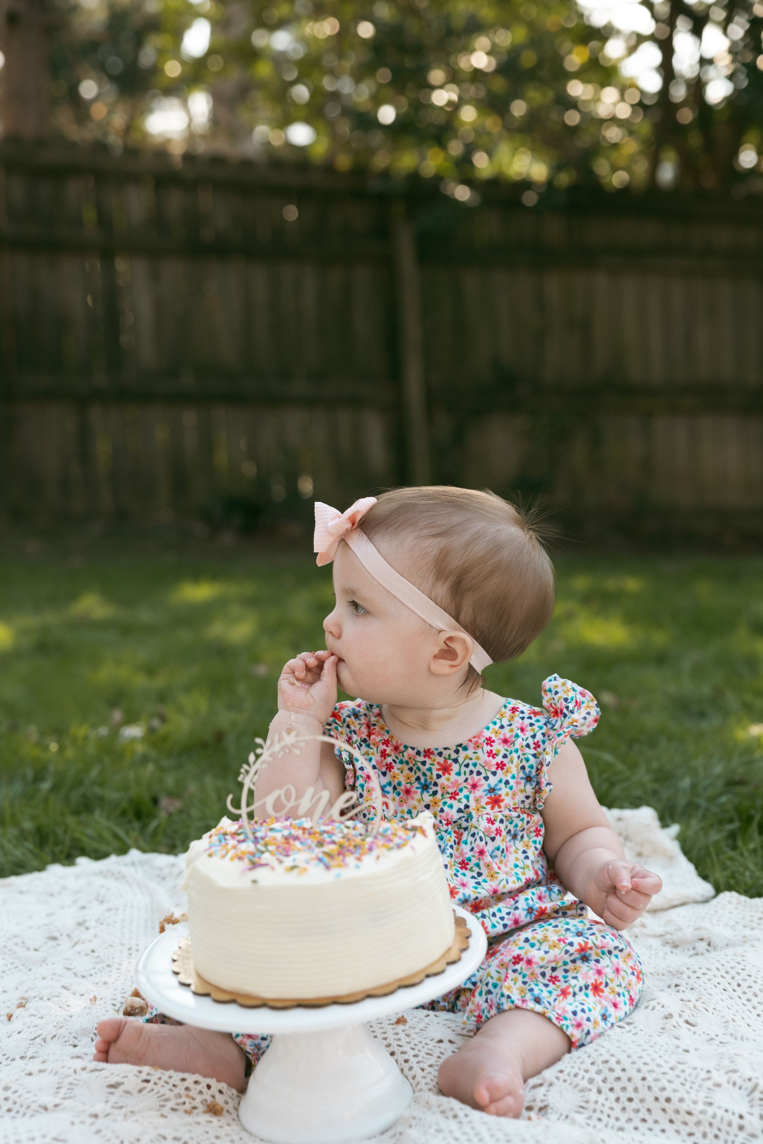Baby girl in floral romper with pink bow headband eating cake at outdoor first birthday smash cake photo session.