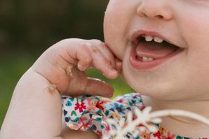 Close-up of a smiling baby with two tiny teeth, chewing on fingers, wearing a floral outfit outdoors.