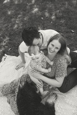 Black and white photo of a happy family with a baby and dog relaxing on a blanket outdoors on grass.