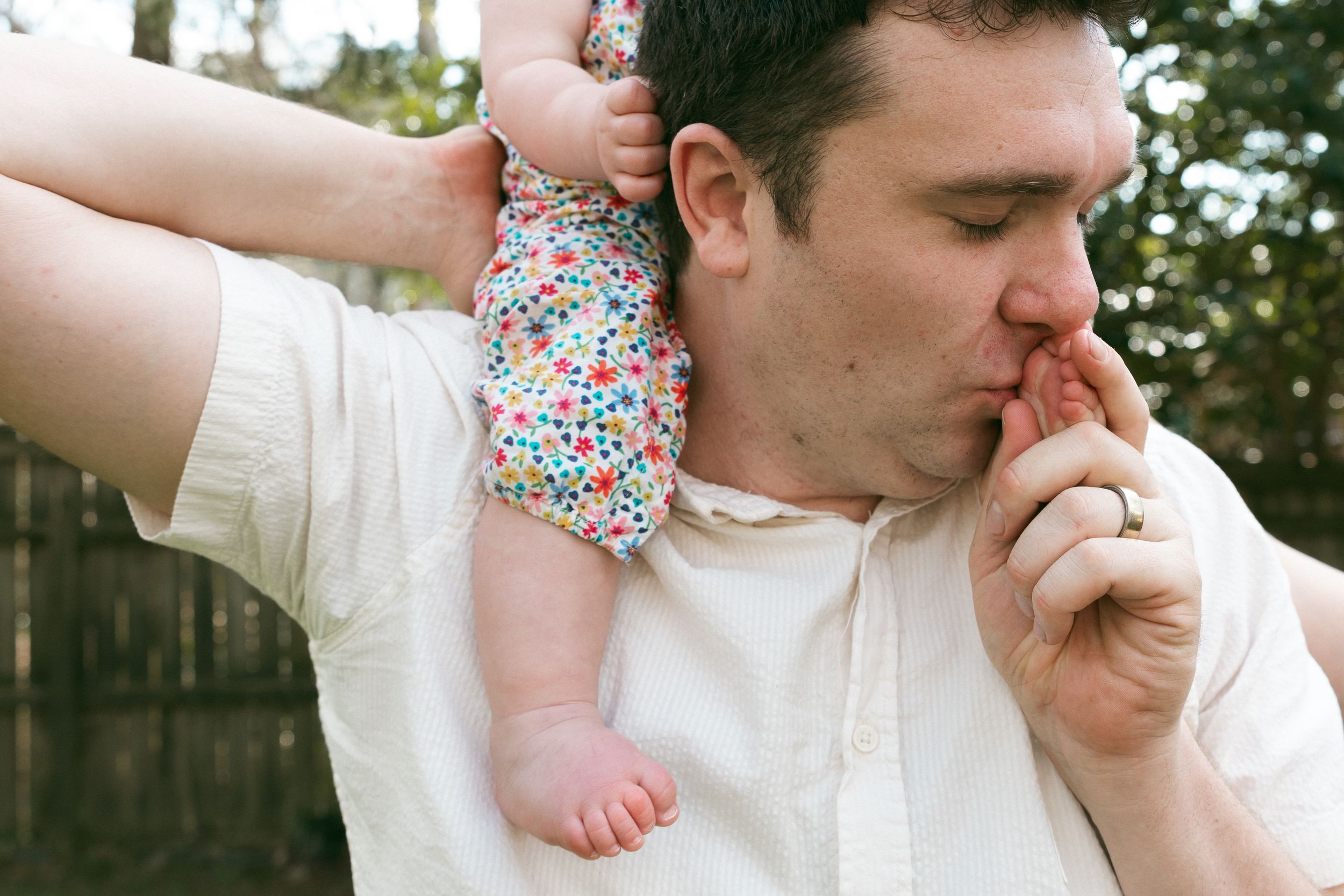A father tenderly kisses his baby's hand while holding the infant on his shoulder outdoors.
