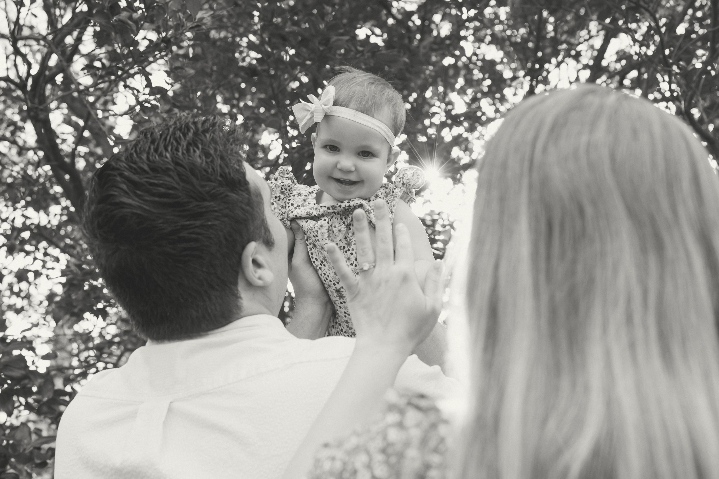 Black and white photo of parents lifting smiling baby girl outdoors under trees.