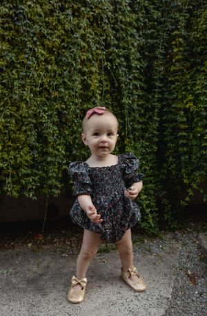 A toddler in a dark floral romper poses against a lush green hedge wall.