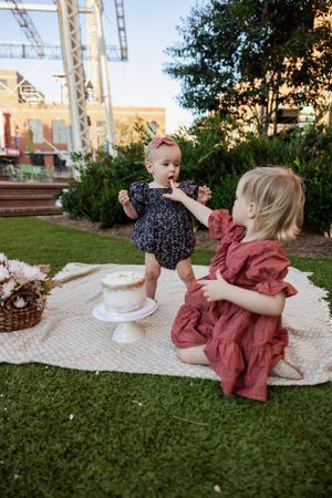 Two children in floral and rust-colored dresses playing during an outdoor cake celebration.