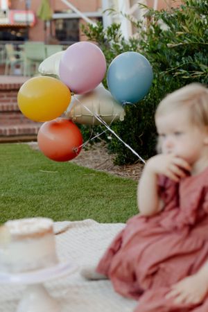 Colorful party balloons against greenery with cake and child in rust dress in foreground.
