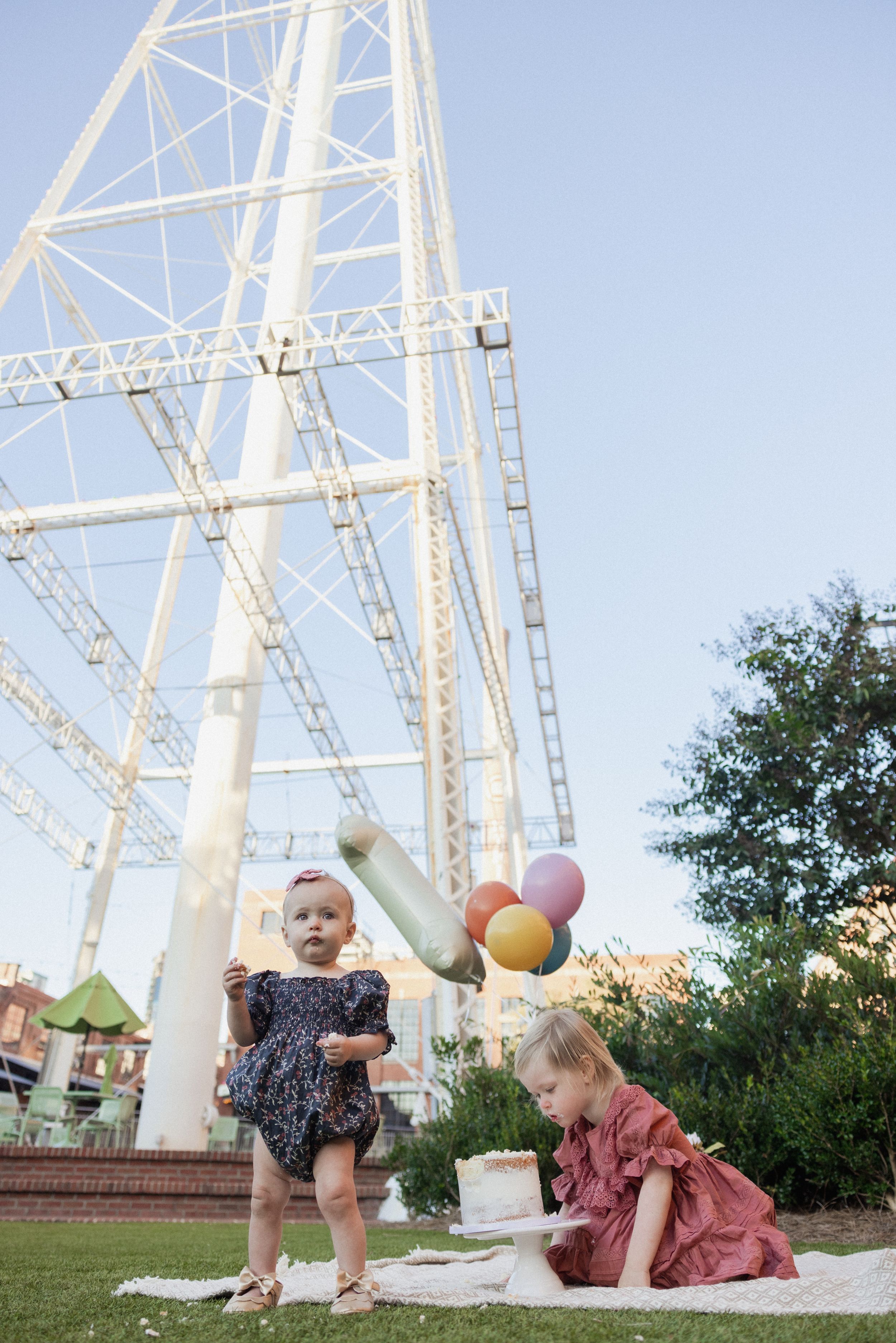 Two children in party dresses playing on a blanket with a ferris wheel structure in the background.