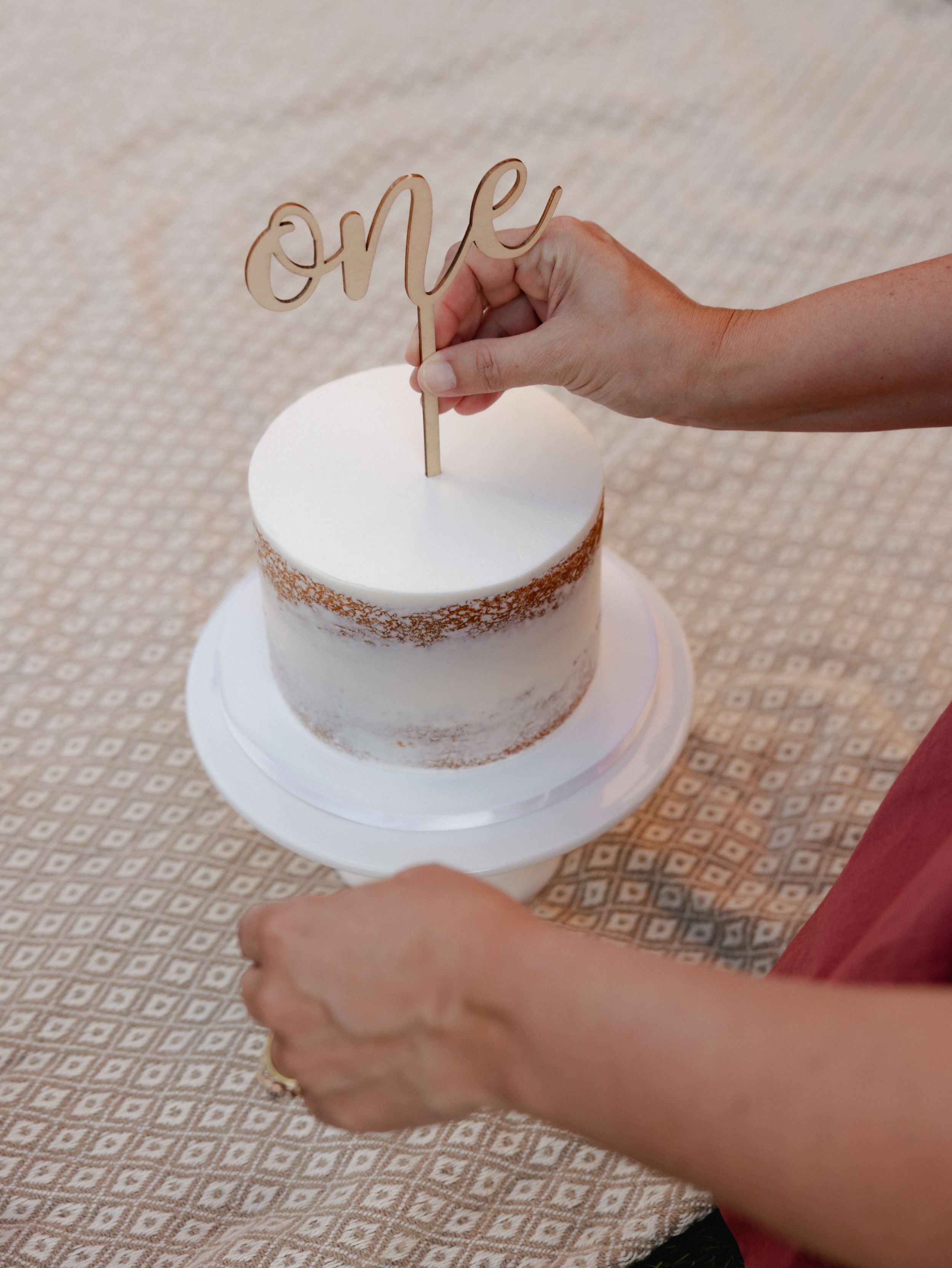 Hands placing a gold 'ONE' cake topper on a white frosted cake.