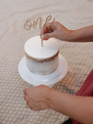 Hands placing a gold 'ONE' cake topper on a white frosted cake.