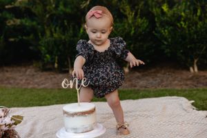 Small child in a dark floral dress standing next to their first birthday cake.