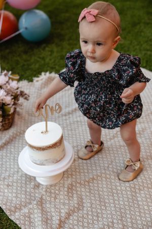 Small child in a floral dress standing next to a white birthday cake with gold decorations.