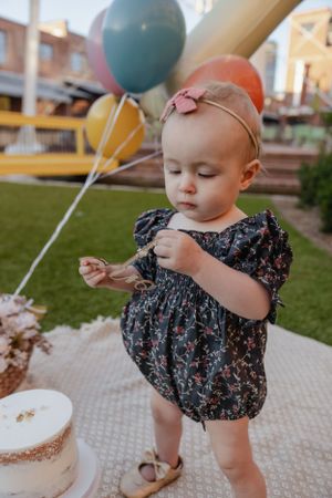Child in a patterned dress examining something while standing near balloons and a cake.