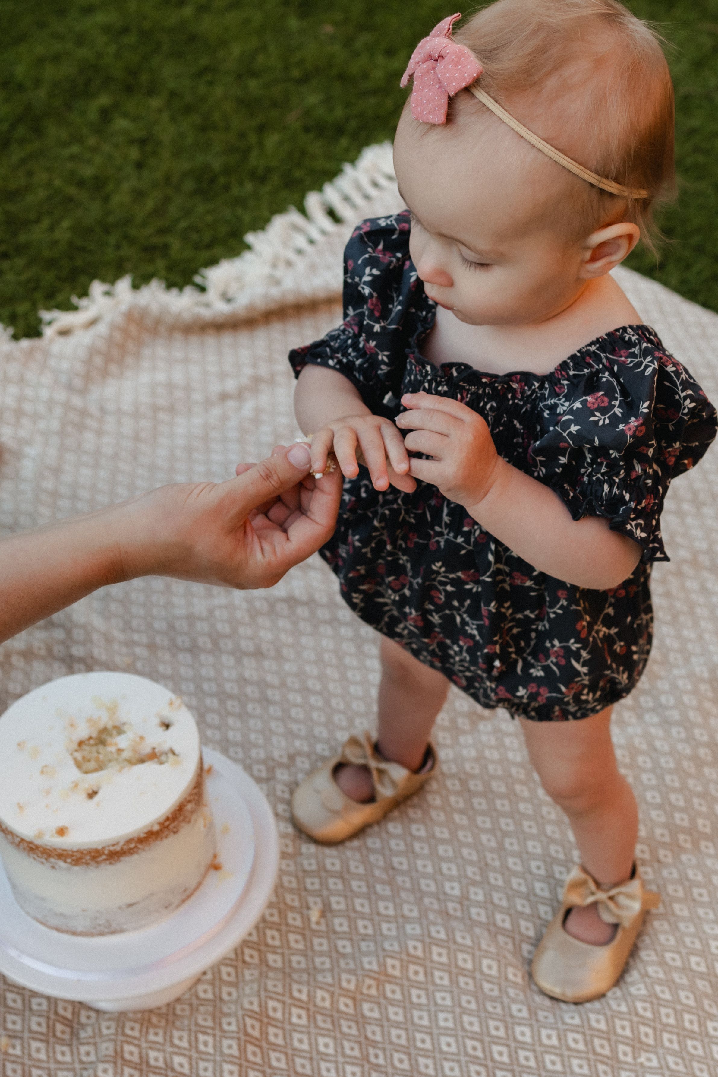 A small child in a floral dress and pink bow reaches for a white cake during an outdoor celebration.
