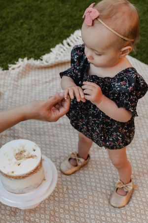 A small child in a floral dress and pink bow reaches for a white cake during an outdoor celebration.
