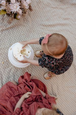 Overhead view of a toddler in dark floral dress touching a white cake on a textured blanket.