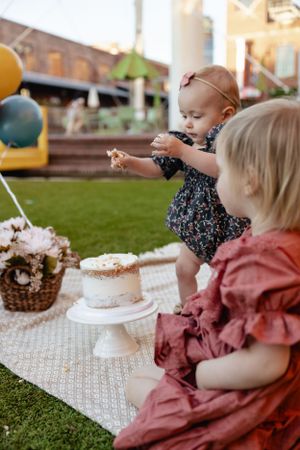 Two small children in dresses sitting on a blanket with cake and balloons at an outdoor party.