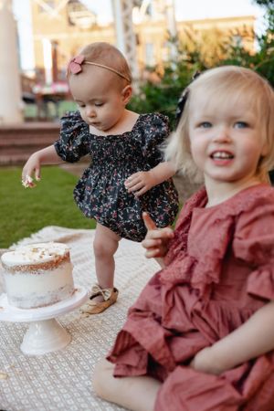 Two small children in patterned dresses enjoying cake time on a picnic blanket outdoors.