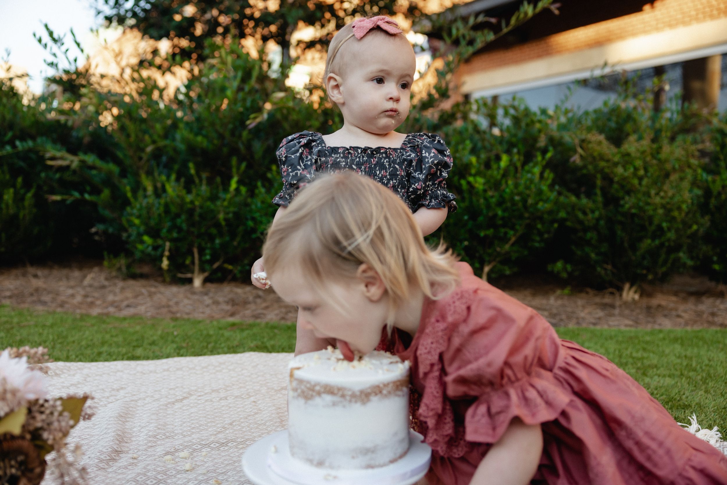 Two children in ruffled dresses exploring a white cake during an outdoor celebration.