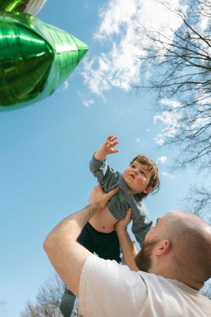 Father lifting toddler toward green balloon against blue sky outdoors.