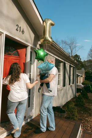Family with balloons entering a gray house with red door, number 21 visible, celebrating a first birthday outdoors.