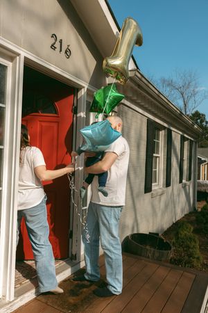 Couple with baby and star balloons entering red door of gray house numbered 216.