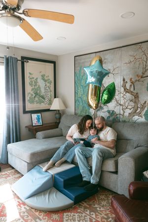 Family with baby sitting on gray sofa with colorful star balloons in cozy decorated living room.