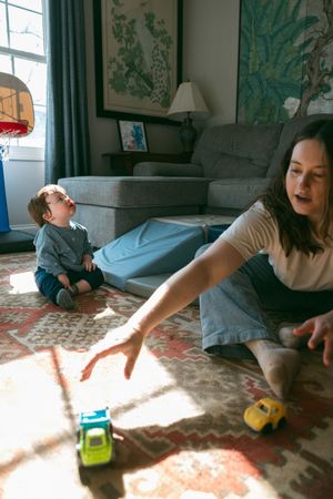 A mother plays with toy cars on a rug while her baby sits nearby in a sunlit living room.