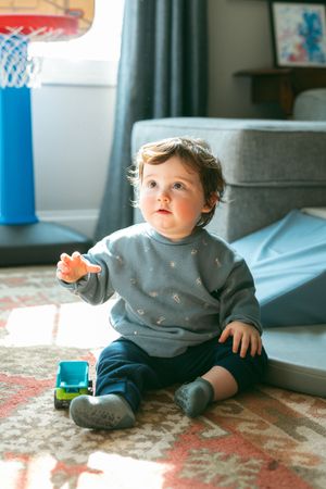 Curious toddler in gray sweatshirt sitting on colorful rug, reaching out while playing with toy truck indoors.