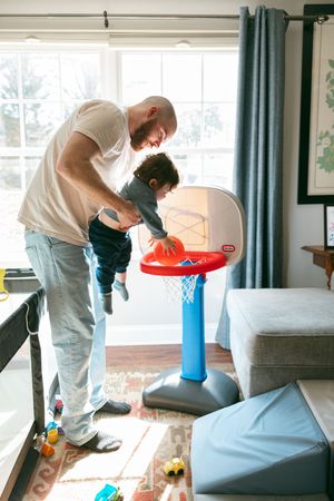 Father lifts toddler to dunk a basketball on a Little Tikes hoop indoors.