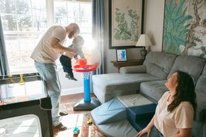 Father lifting baby toward toy basketball hoop while mother watches in cozy living room.