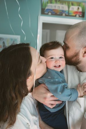 Parents kissing their smiling baby on both cheeks in a cozy teal nursery room.