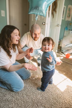 Happy toddler standing with parents holding a blue star balloon in a sunny bedroom during first birthday celebration.