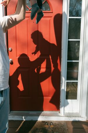 Shadows of parent lifting child cast on red front door in sunlight.