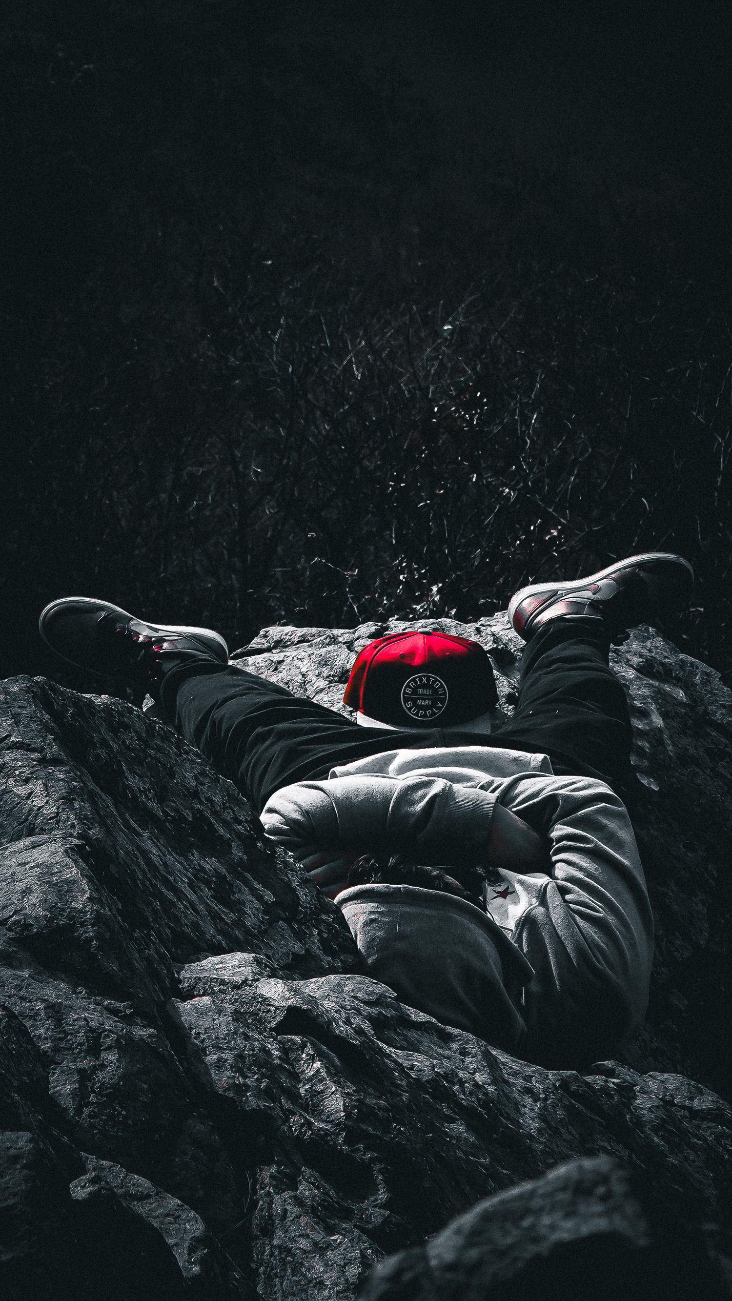 Person lying on dark rocks wearing a red cap captured in dramatic monochromatic style.