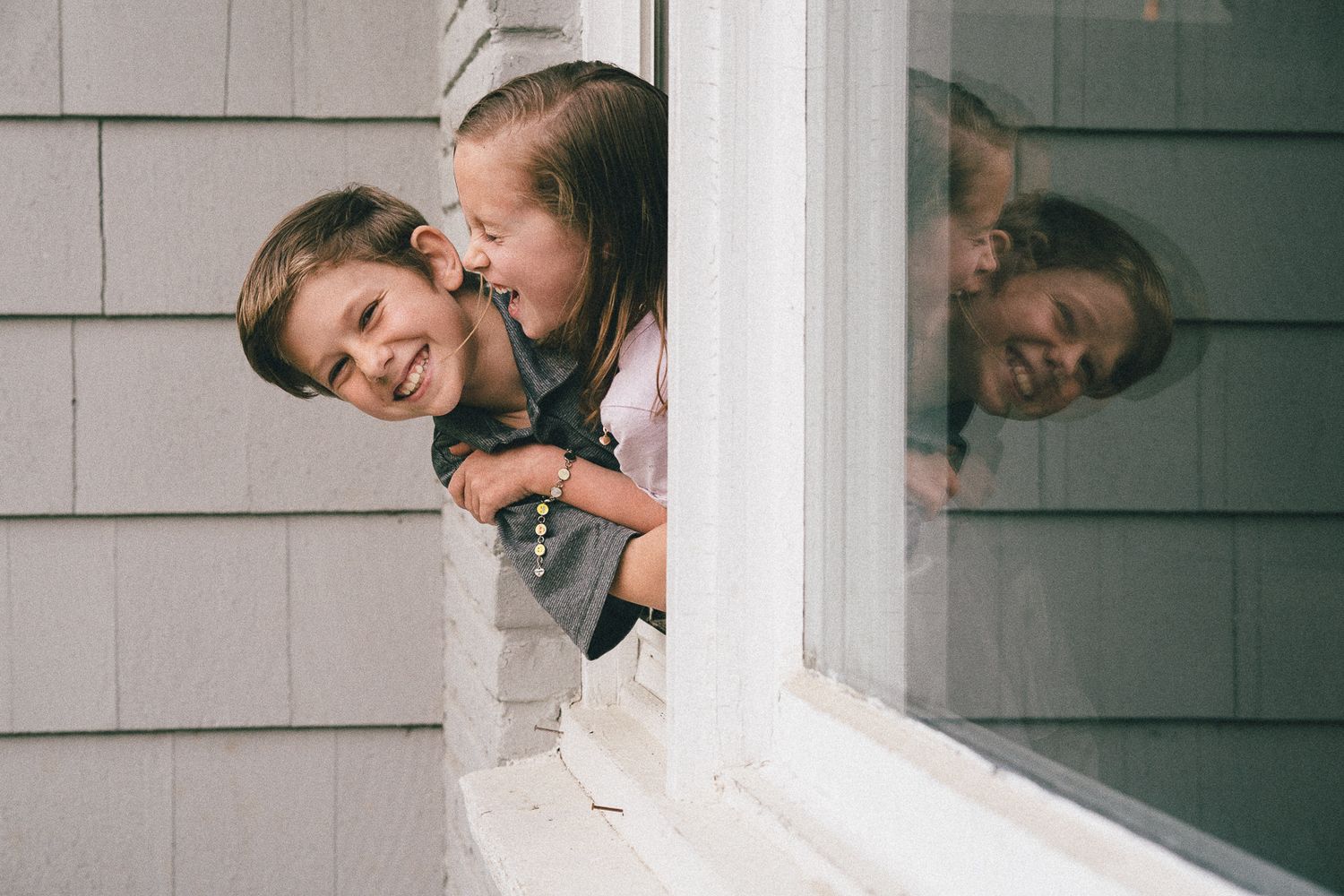 Two people playfully peeking out of a white window frame with their reflection visible.