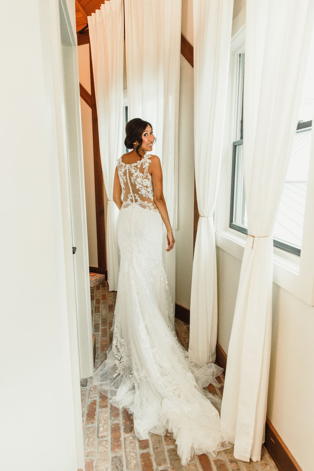 Bride in lace wedding gown posing by white curtained windows.