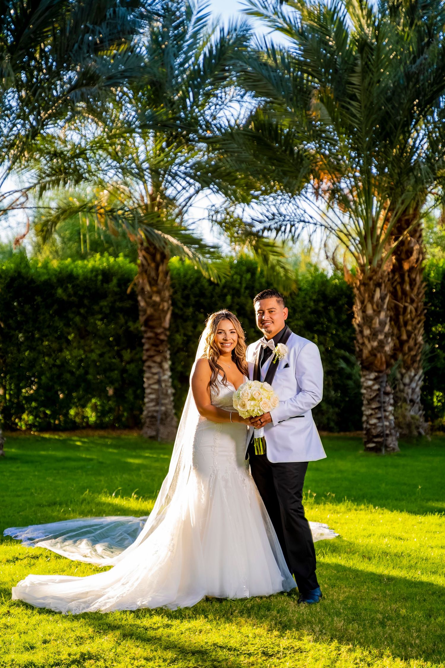 Newlyweds pose amid palm trees on a sun-drenched lawn during their wedding photoshoot.
