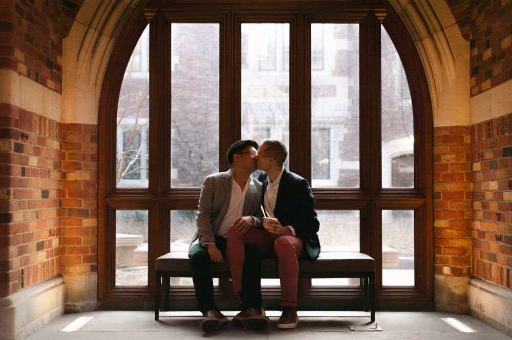 Two people share a romantic kiss while seated on a bench in front of an arched window in a brick building.