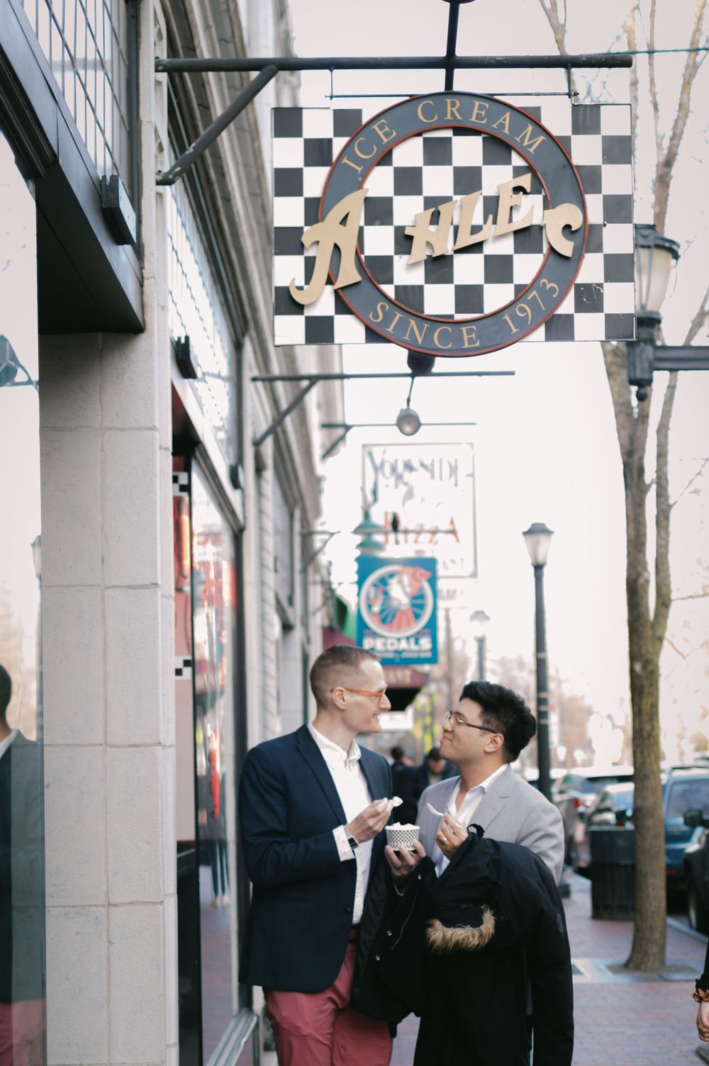 Two men share a lighthearted conversation outside a vintage ice cream shop during their engagement photo session.