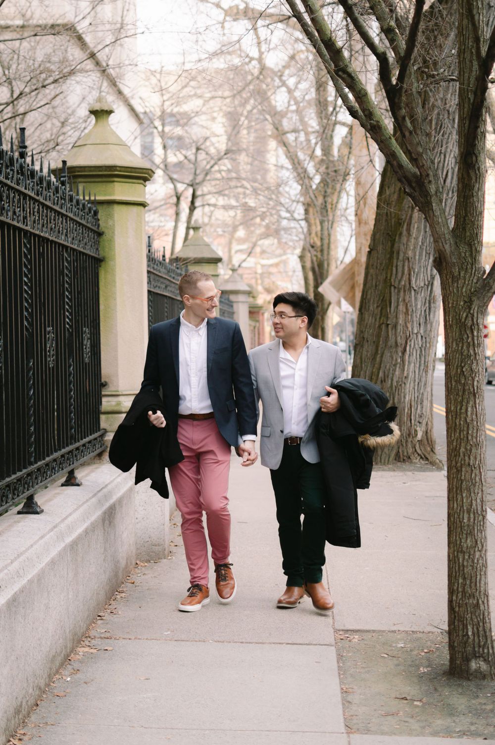 Two men walk hand-in-hand along a city sidewalk lined with iron fencing during their engagement photo session.