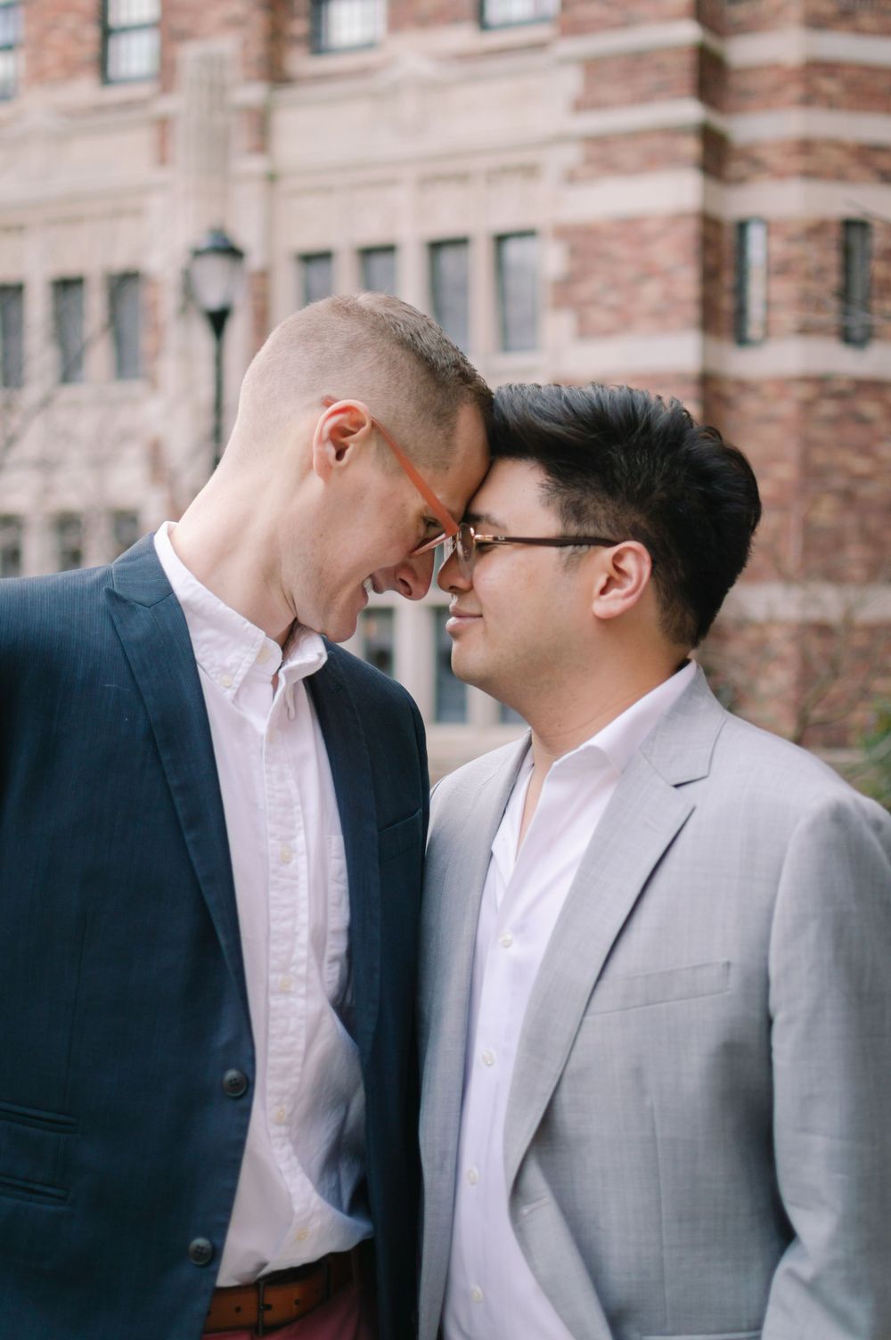 Two men in suits share a tender forehead-to-forehead moment during their engagement session near a brick building.
