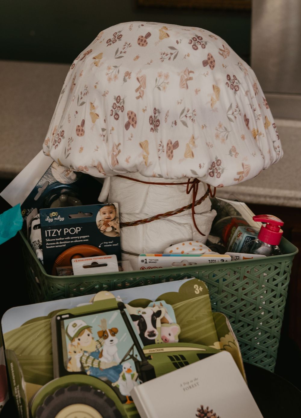 Baby shower gift basket filled with Itzy Pop pacifier, muslin blanket, and baby essentials in green container.