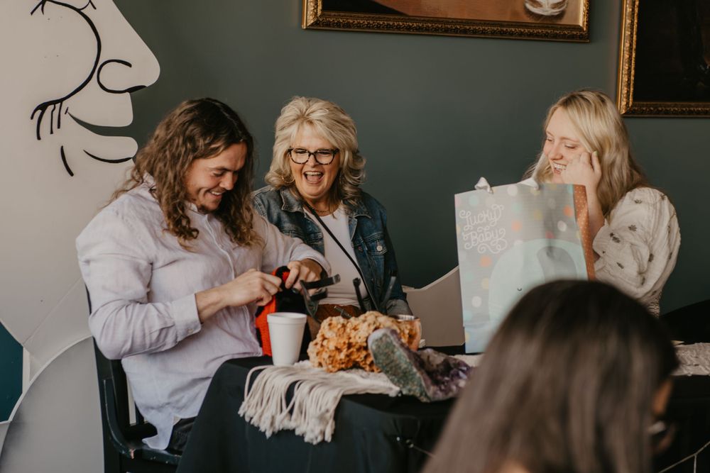 Three women laughing and opening gifts together at baby shower seated near crescent moon decoration.