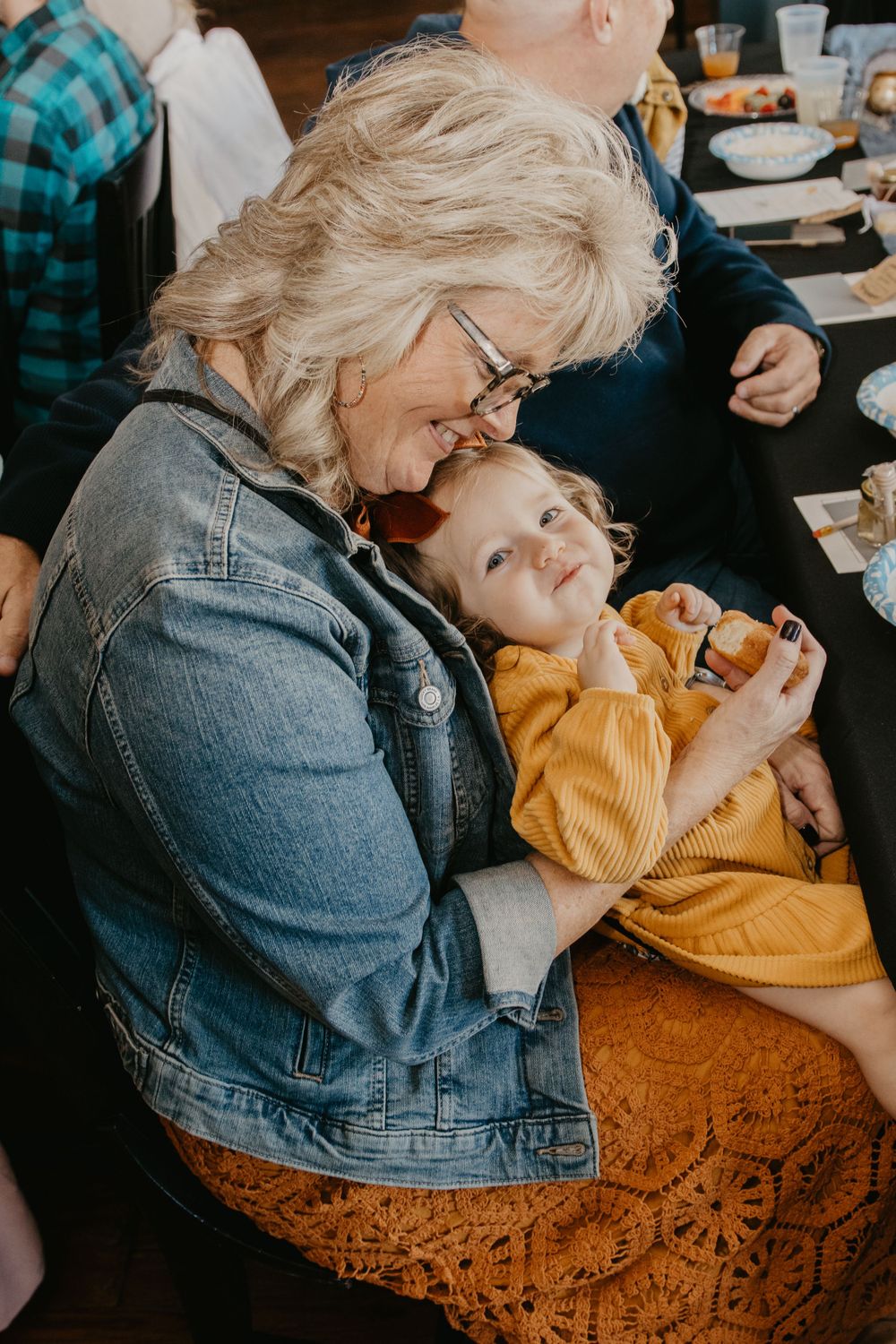 Grandmother with glasses in denim jacket lovingly holding smiling toddler in yellow outfit at baby shower.