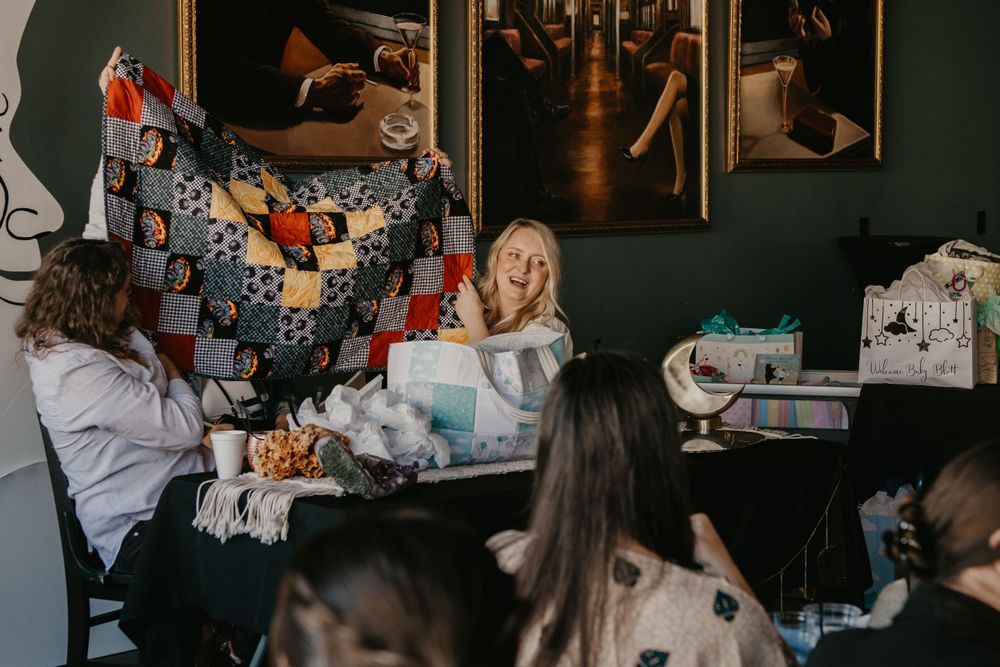 Woman holding up a colorful patchwork quilt at an indoor baby shower gathering with guests seated around tables.