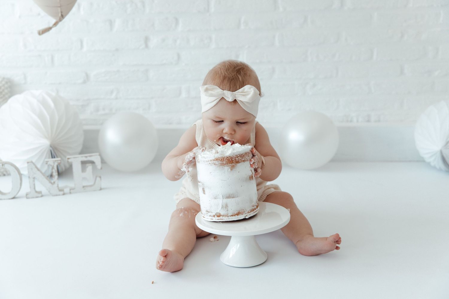 Baby wearing white bow headband sits with small white cake on pedestal for first birthday photoshoot.