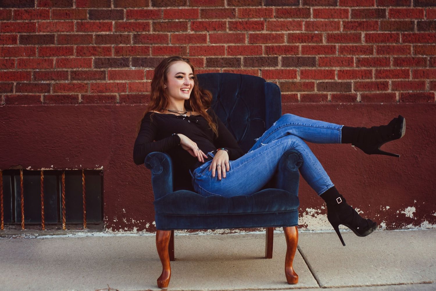 A woman in blue jeans lounges playfully on a navy blue armchair against a brick wall.