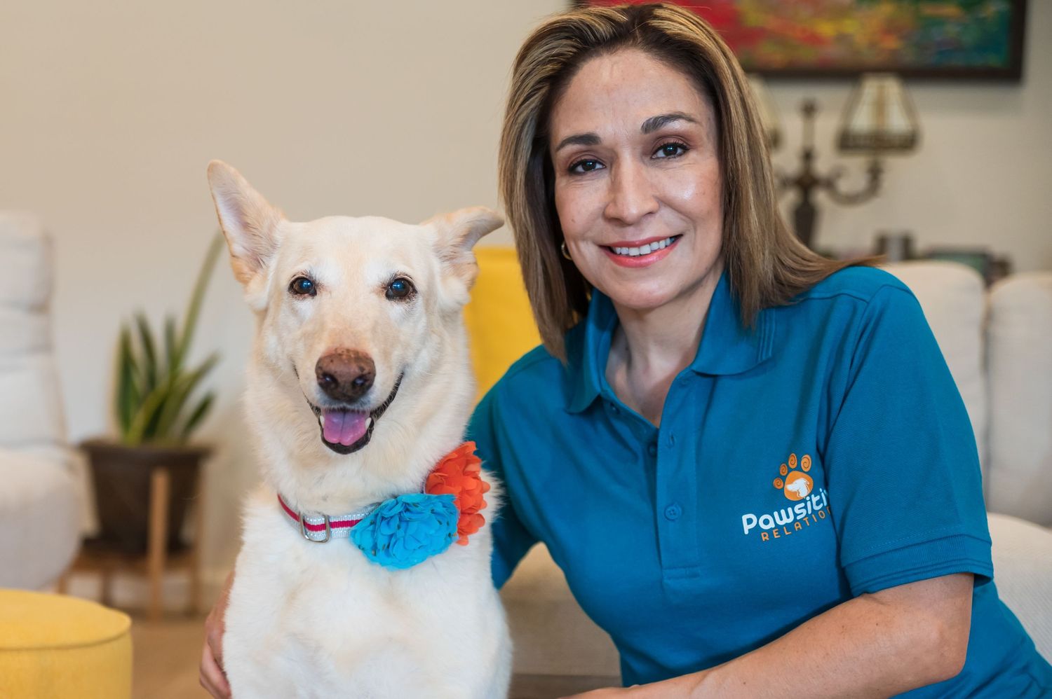 A happy white dog with a blue collar flower poses with a pet care professional.