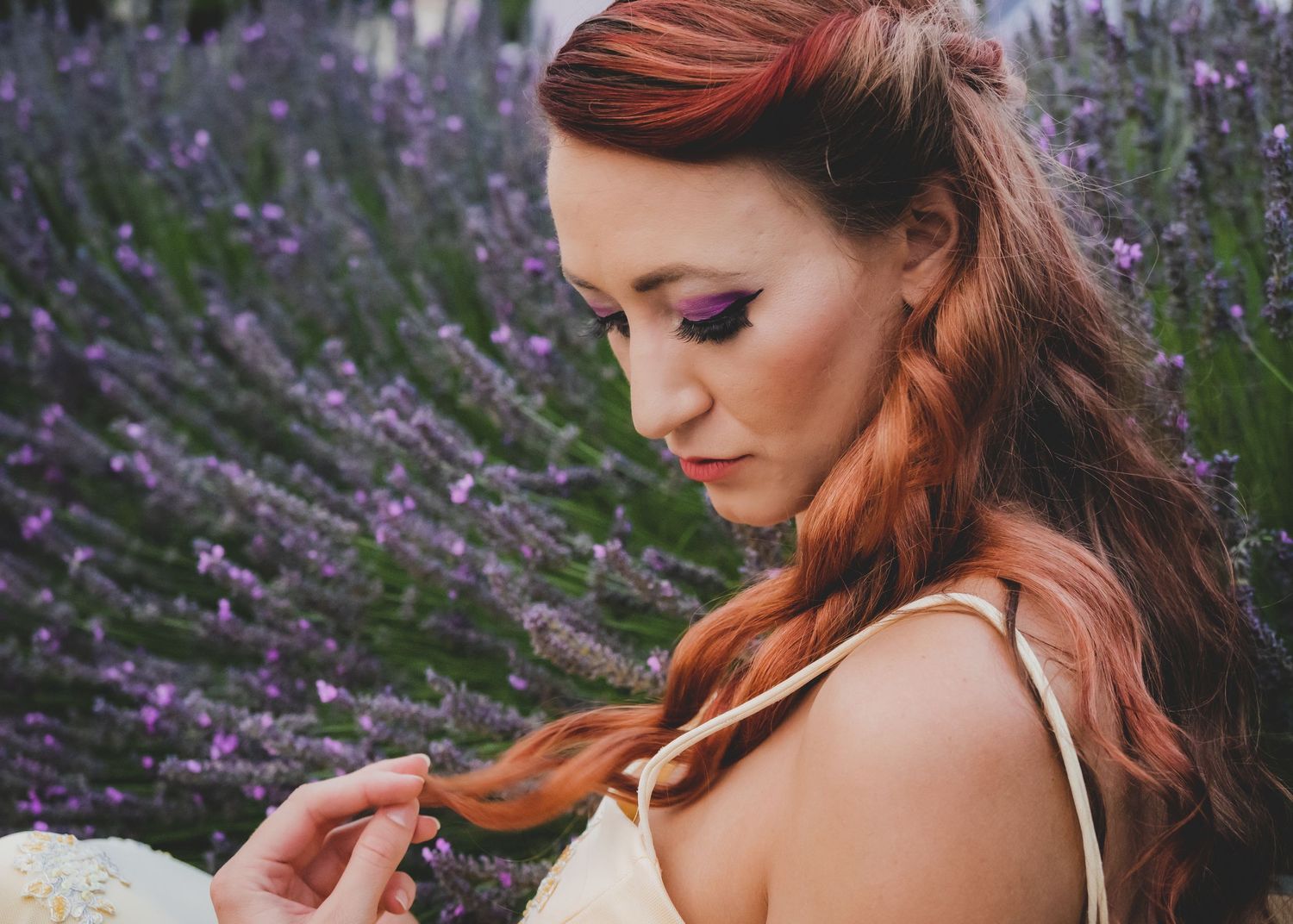 Woman with red braided hair and purple eye makeup among blooming lavender flowers in a field.