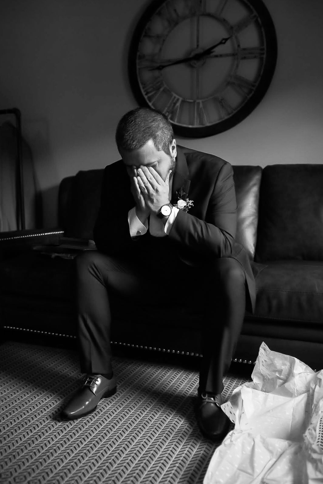 Emotional groom in suit covering face while sitting on couch beneath vintage clock.