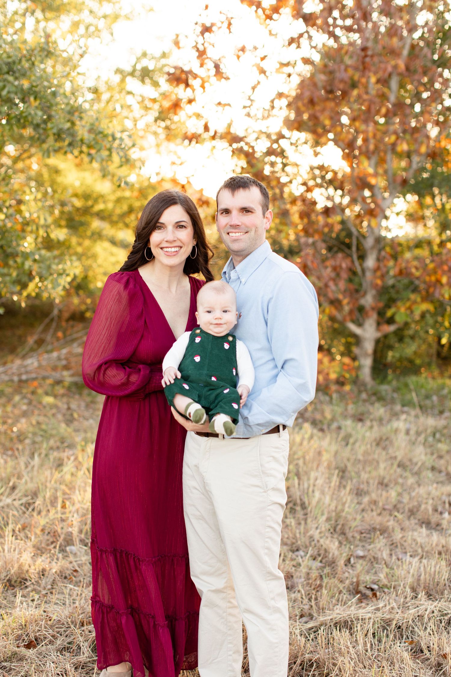 Family portrait in autumn setting with burgundy dress and green overalls against golden foliage background.