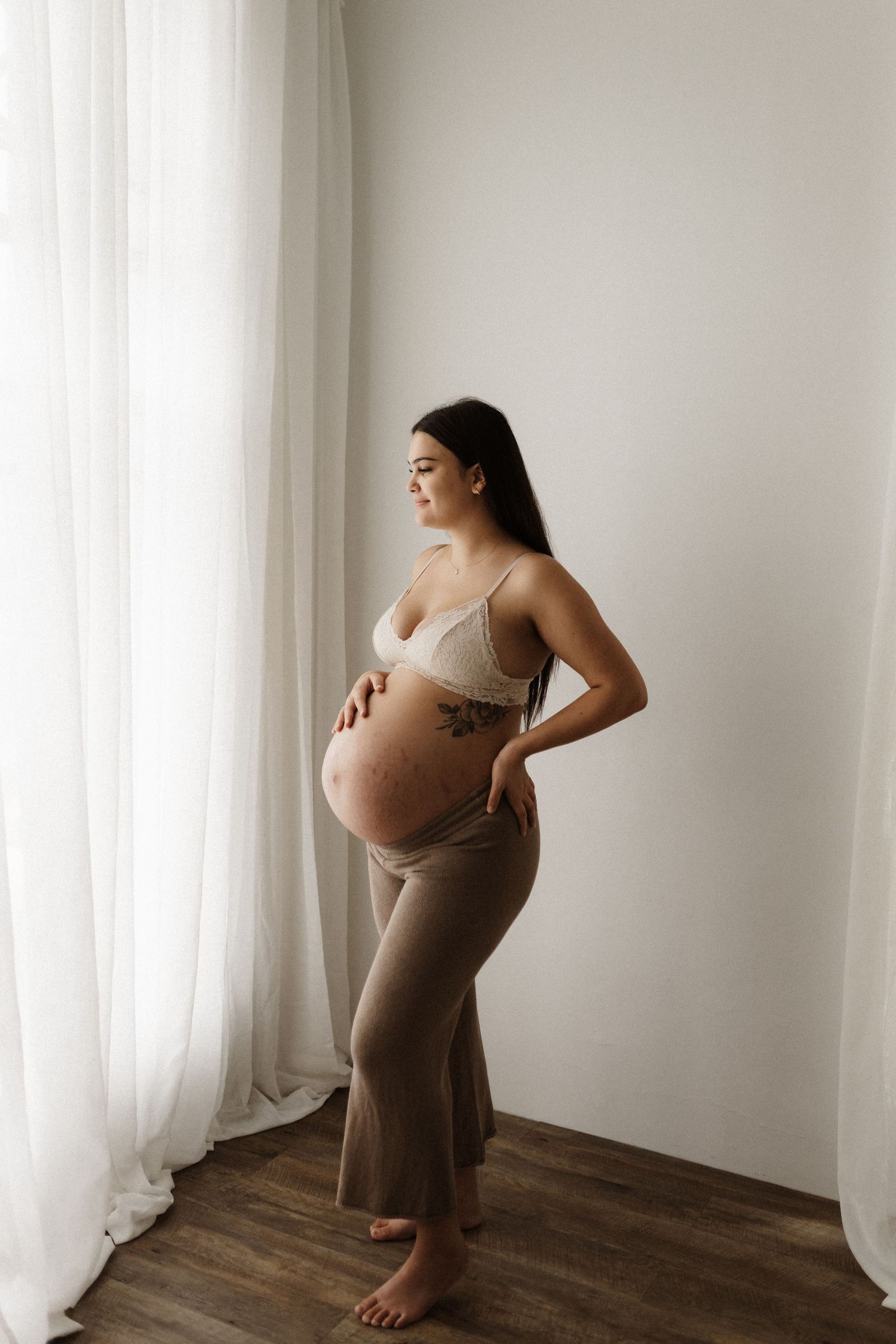 Side view of pregnant person in brown dress standing by window in minimalist setting.